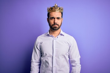 Young handsome man with beard wearing golden crown of king over purple background skeptic and nervous, frowning upset because of problem. Negative person.