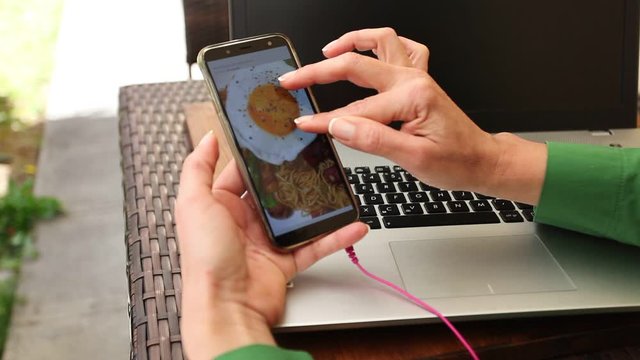 Close Up Of Woman Hand Scrolling Smartphone Screen On Which A Cooking Video Passes. The Arms Are Resting On A Laptop