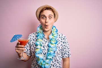 Young redhead tourist man on vacation wearing hat and hawaiian lei drinking cocktail scared and...