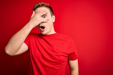 Young handsome redhead man wearing casual t-shirt over isolated red background peeking in shock covering face and eyes with hand, looking through fingers with embarrassed expression.