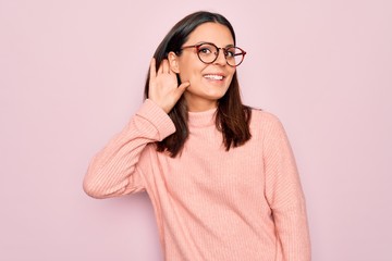 Young beautiful brunette woman wearing casual sweater and glasses over pink background smiling with hand over ear listening an hearing to rumor or gossip. Deafness concept.