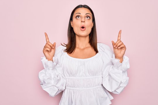 Young beautiful brunette woman wearing casual white dress standing over pink background amazed and surprised looking up and pointing with fingers and raised arms.