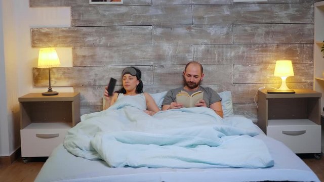 Beautiful wife turing the room lights from phone app before going to sleep. Husband reading a book.