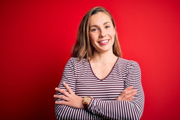 Obraz premium Young beautiful blonde woman wearing casual striped t-shirt over isolated red background happy face smiling with crossed arms looking at the camera. Positive person.