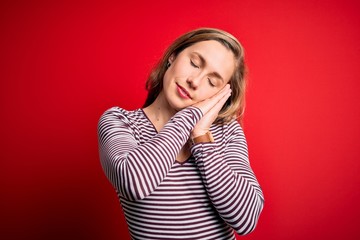 Young beautiful blonde woman wearing casual striped t-shirt over isolated red background sleeping tired dreaming and posing with hands together while smiling with closed eyes.