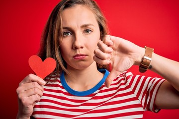 Young beautiful blonde woman holding paper heart standing over isolated red background with angry face, negative sign showing dislike with thumbs down, rejection concept