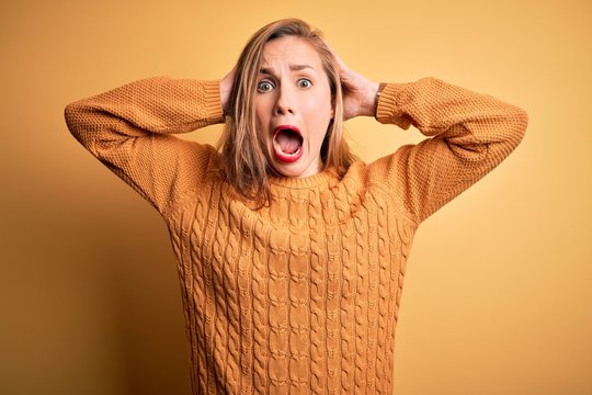 Young beautiful blonde woman wearing casual sweater standing over yellow background Crazy and scared with hands on head, afraid and surprised of shock with open mouth