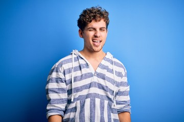 Young blond handsome man with curly hair wearing casual striped sweatshirt winking looking at the camera with sexy expression, cheerful and happy face.