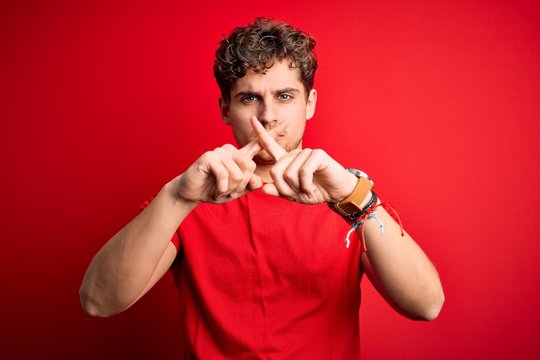 Young blond handsome man with curly hair wearing casual t-shirt over red background Rejection expression crossing fingers doing negative sign