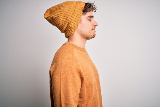 Young blond handsome man with curly hair wearing casual sweater and wool hat looking to side, relax profile pose with natural face with confident smile.