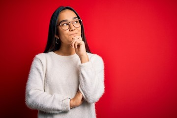 Young beautiful asian woman wearing casual sweater and glasses over red background with hand on chin thinking about question, pensive expression. Smiling and thoughtful face. Doubt concept.