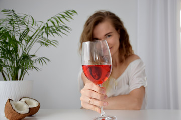 Beautiful girl with curly hair with naked shoulder toasts with red drink near palm tree in white pot and coconut