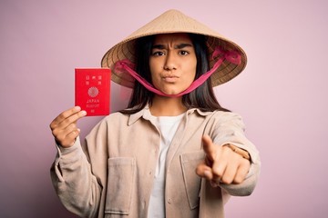 Young thai woman wearing traditional conical asian hat holding japan japanese passport pointing with finger to the camera and to you, hand sign, positive and confident gesture from the front