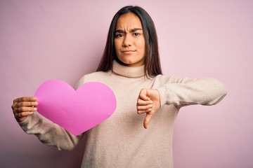 Young beautiful asian woman holding pink heart standing over isolated background with angry face, negative sign showing dislike with thumbs down, rejection concept