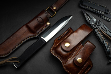 Hunting knife and multitool on a dark background. Brown leather cases near the knife and multitool. Postcard concept for men.