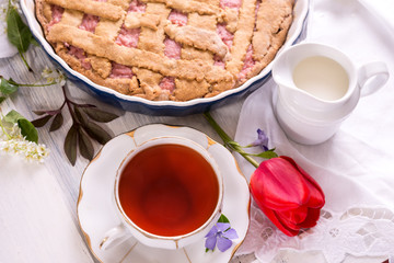 White porcelain cup of tea, and fresh-baked pie. English breakfast still-life with drink and treats red tulip flower and white tablecloth.
