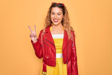 Young beautiful blonde woman pin-up with blue eyes wearing red sunglasses and jacket smiling with happy face winking at the camera doing victory sign. Number two.