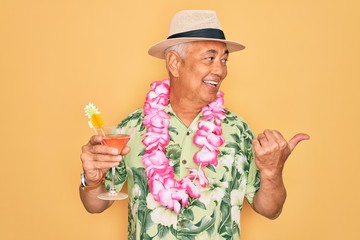 Middle age senior grey-haired man wearing summer hat and hawaiian lei drinking a cocktail pointing...
