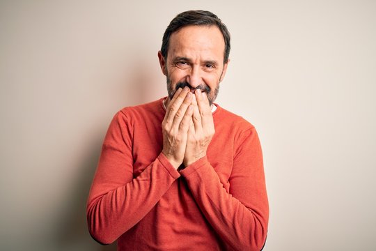 Middle age hoary man wearing casual orange sweater standing over isolated white background laughing and embarrassed giggle covering mouth with hands, gossip and scandal concept
