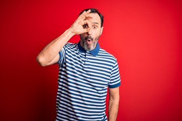 Middle age hoary man wearing casual striped polo standing over isolated red background doing ok gesture shocked with surprised face, eye looking through fingers. Unbelieving expression.