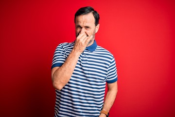 Middle age hoary man wearing casual striped polo standing over isolated red background smelling something stinky and disgusting, intolerable smell, holding breath with fingers on nose. Bad smell