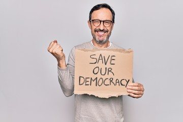 Middle age man holding banner with saver our democracy message over white background screaming proud, celebrating victory and success very excited with raised arm
