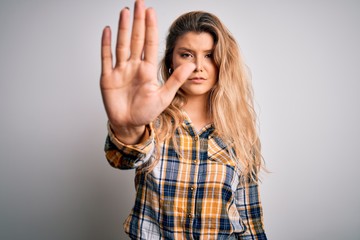 Young beautiful blonde woman wearing casual shirt standing over isolated white background doing stop sing with palm of the hand. Warning expression with negative and serious gesture on the face.