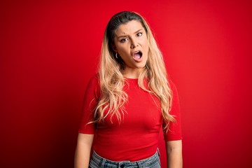 Young beautiful blonde woman wearing casual t-shirt standing over isolated red background In shock...