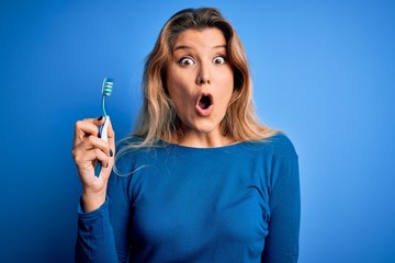 Young beautiful blonde woman using teeth brush over isolated blue background scared in shock with a...