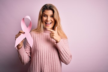 Young beautiful blonde woman asking for support cancer holding pink ribbon symbol very happy pointing with hand and finger