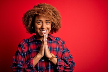 Young beautiful African American afro woman with curly hair wearing casual shirt praying with hands together asking for forgiveness smiling confident.