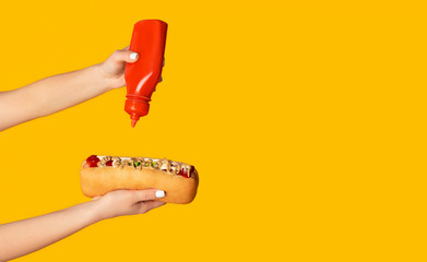 Close up of young woman squeezing ketchup onto hot dog against orange background, copy space....