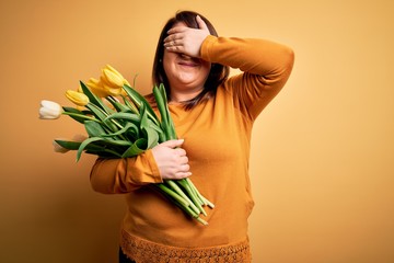 Beautiful plus size woman holding romantic bouquet of natural tulips flowers over yellow background smiling and laughing with hand on face covering eyes for surprise. Blind concept.