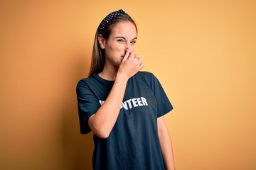 Young beautiful woman wearing volunteer t-shirt doing volunteering over yellow background smelling...