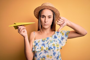 Young tourist woman on vacation wearing summer hat holding hand fan over yellow background with angry face, negative sign showing dislike with thumbs down, rejection concept