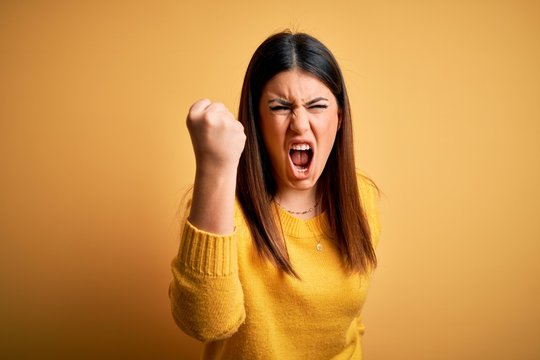 Young beautiful woman wearing casual sweater over yellow isolated background angry and mad raising fist frustrated and furious while shouting with anger. Rage and aggressive concept.