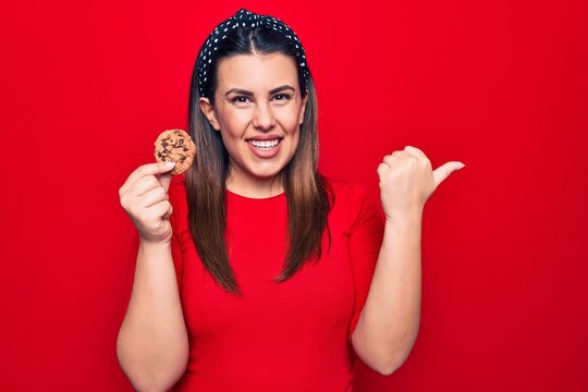 Young beautiful brunette woman holding sweet chocolate cookie over isolated red background pointing thumb up to the side smiling happy with open mouth