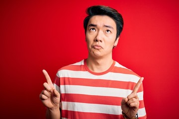 Young handsome chinese man wearing casual striped t-shirt standing over red background Pointing up looking sad and upset, indicating direction with fingers, unhappy and depressed.