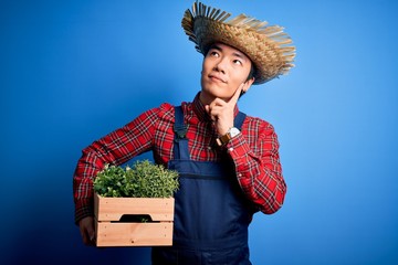 Young handsome chinese farmer man wearing apron and straw hat holding box with plants serious face...