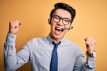 Young handsome chinese call center agent man wering glasses using headset celebrating surprised and amazed for success with arms raised and open eyes. Winner concept.