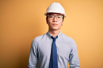 Young handsome chinese architect man wearing safety helmet and tie over yellow background with serious expression on face. Simple and natural looking at the camera.