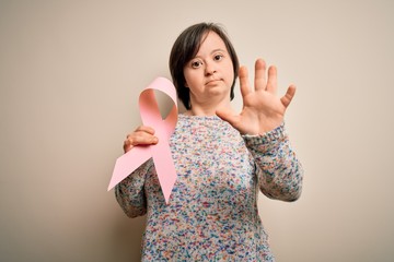 Young down syndrome woman holding breast cancer awareness pink ribbon with open hand doing stop...