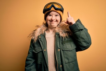 Young down syndrome woman wearing ski coat and glasses for winter weather pointing finger up with successful idea. Exited and happy. Number one.