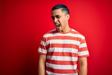 Young brazilian man wearing casual striped t-shirt standing over isolated red background winking...