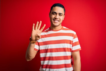 Young brazilian man wearing casual striped t-shirt standing over isolated red background showing and pointing up with fingers number four while smiling confident and happy.