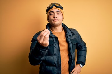 Young brazilian skier man wearing snow sportswear and ski goggles over yellow background Doing Italian gesture with hand and fingers confident expression