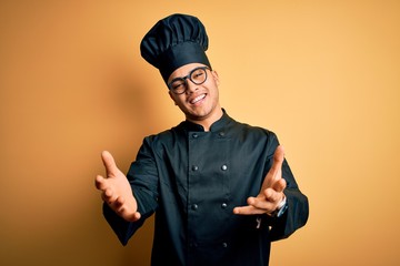 Young brazilian chef man wearing cooker uniform and hat over isolated yellow background smiling cheerful offering hands giving assistance and acceptance.