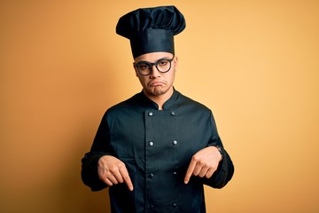 Young brazilian chef man wearing cooker uniform and hat over isolated yellow background Pointing down looking sad and upset, indicating direction with fingers, unhappy and depressed.