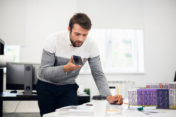 A young architect working on his construction model while talking to someone over the phone using the speaker mode