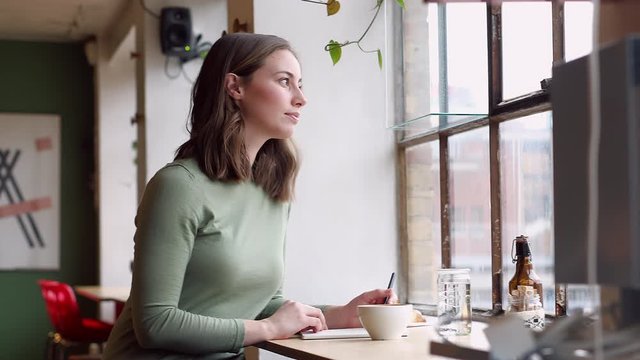 Smiling Young Woman Sitting At A Table At A Cafe Thinking And Writing Notes While Looking Outside The Window Getting Inspired. Freelance, Student, Concept